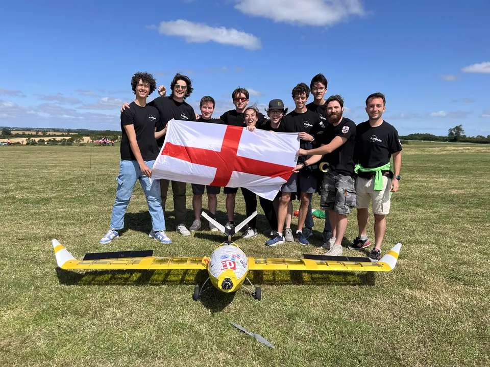 La squadra in maglietta nera tiene la bandiera inglese sul prato del campo di gara; davanti, per terra, il drone giallo Panissa